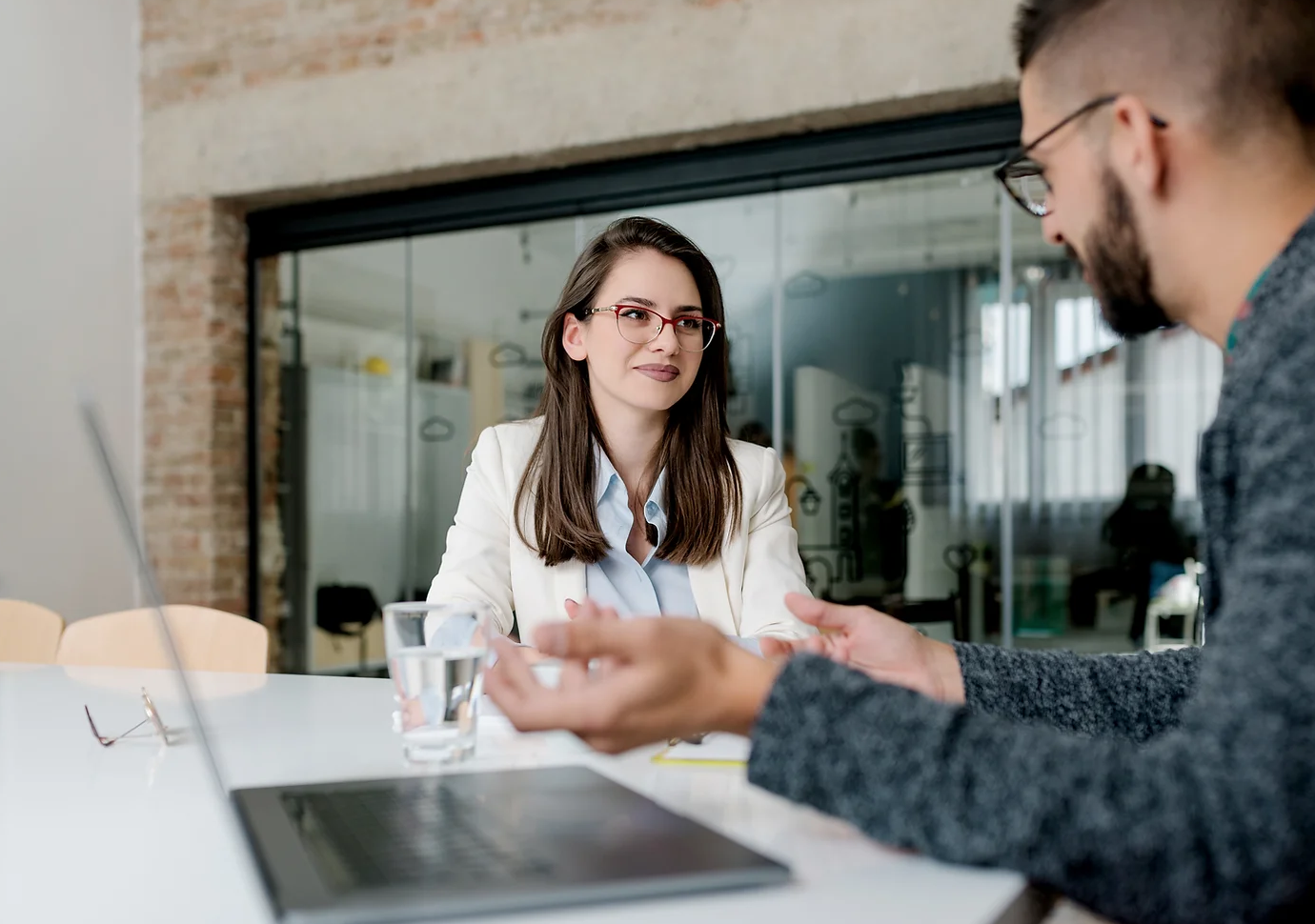 Business meeting with a woman in a white blazer and red glasses and a man with a beard and glasses.
