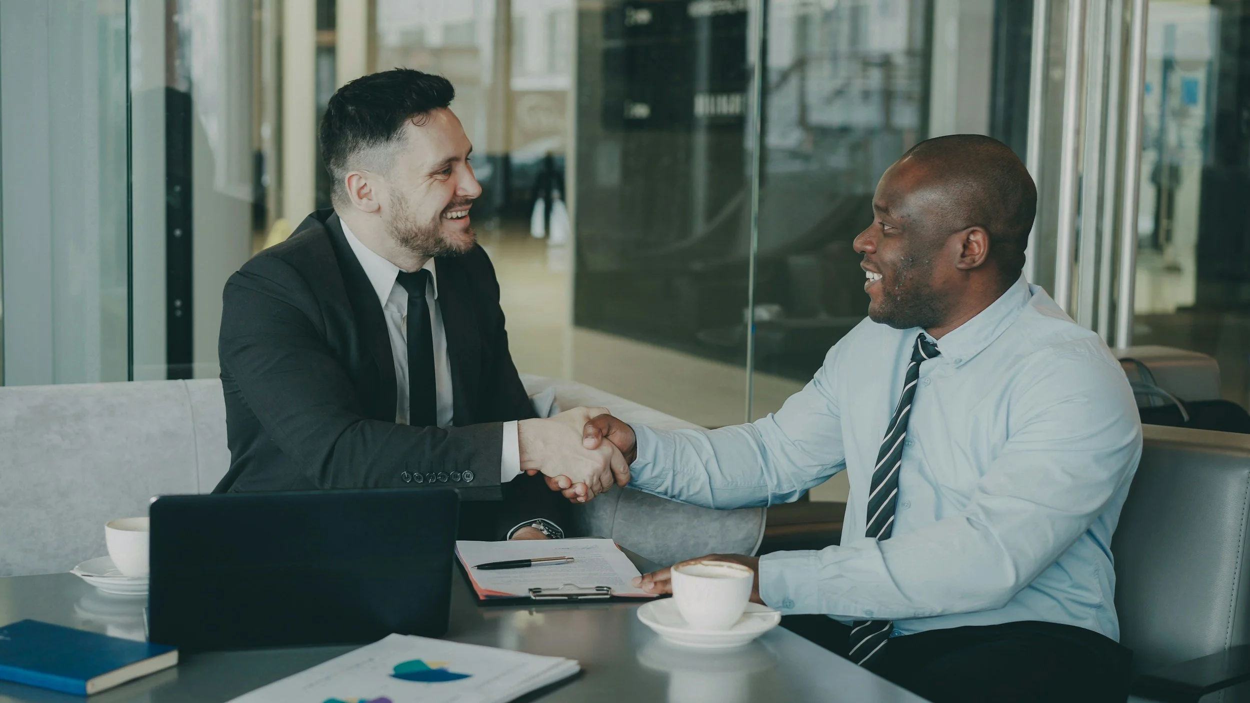 Two businessmen shaking hands over a table with documents and coffee cups.