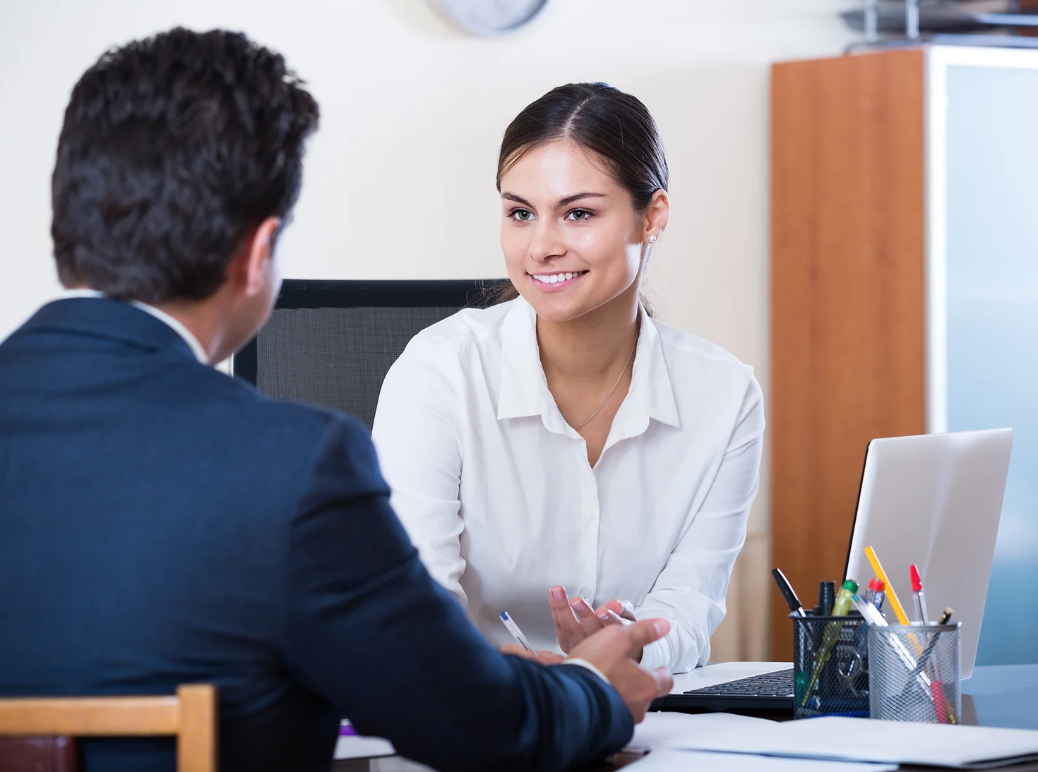 Two professionals in an office setting, a man in a suit and a woman in a white blouse, talking across a desk.