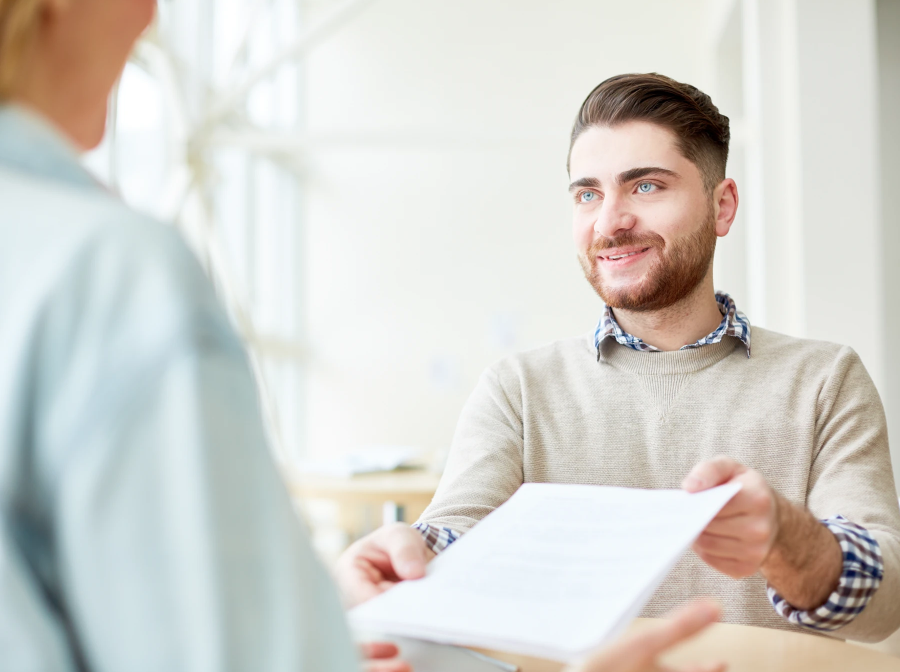 Man in beige sweater smiling and handing a document across a table to someone off-camera.