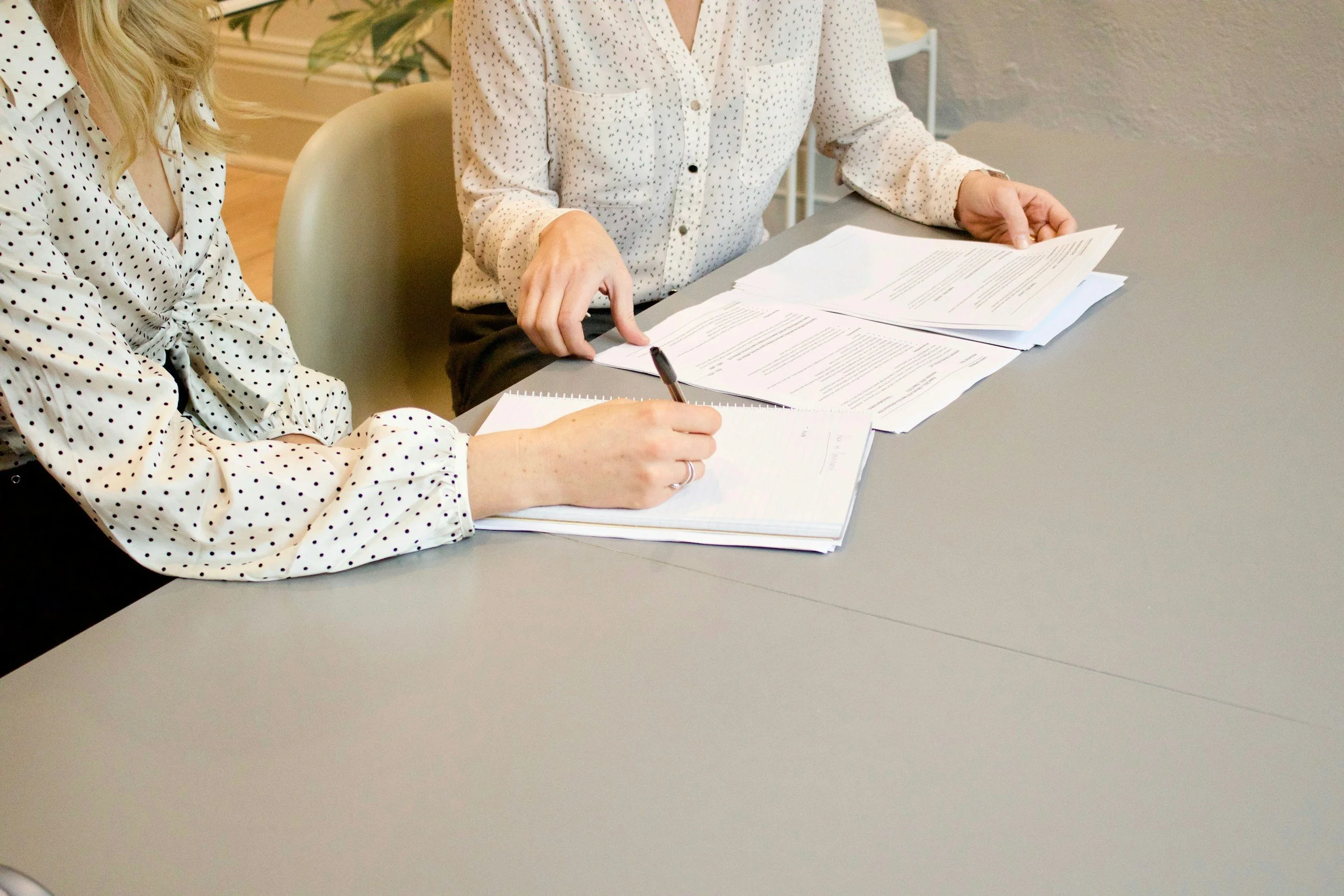 Two women in polka dot shirts review documents at a table, one writing in a notebook.