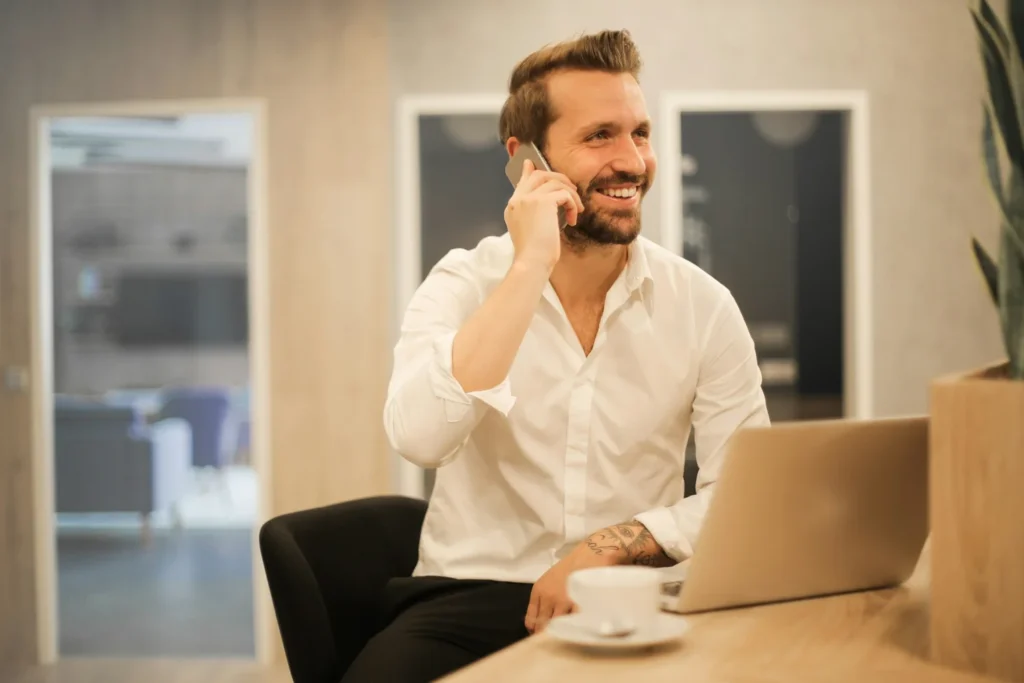 Man in white shirt on phone with laptop and coffee, smiling in office.