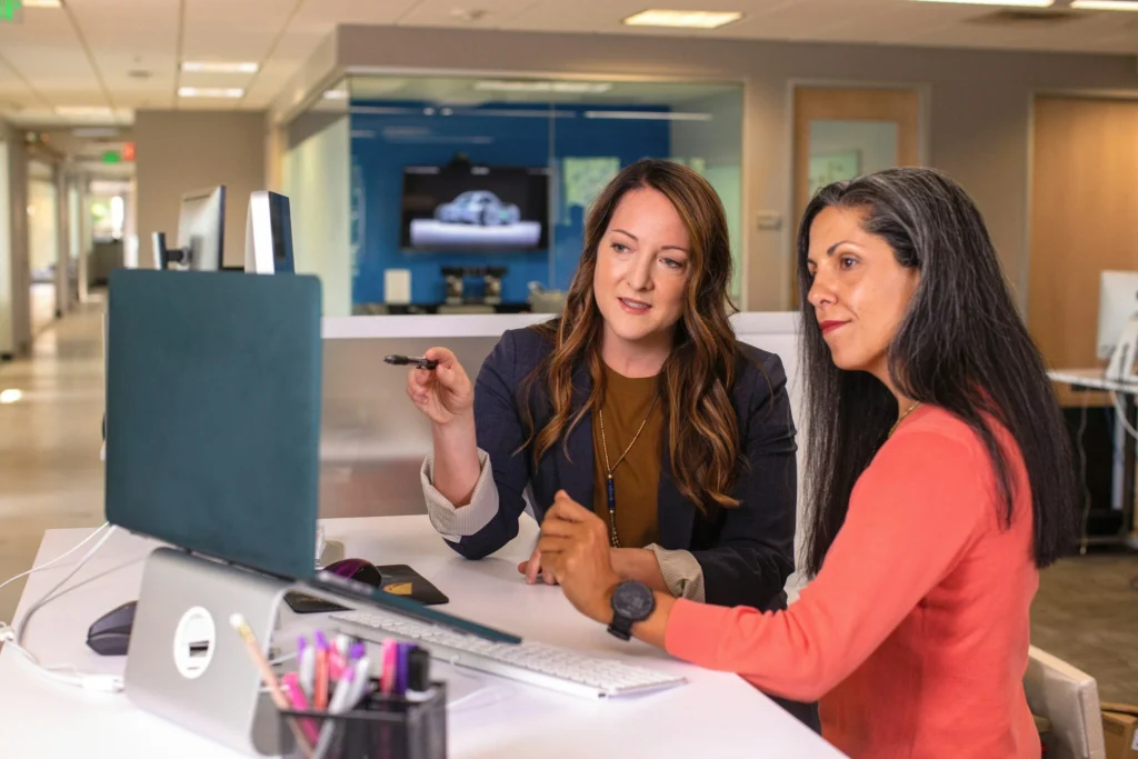 Two women collaborate at a desk with a computer and monitor in a modern office.