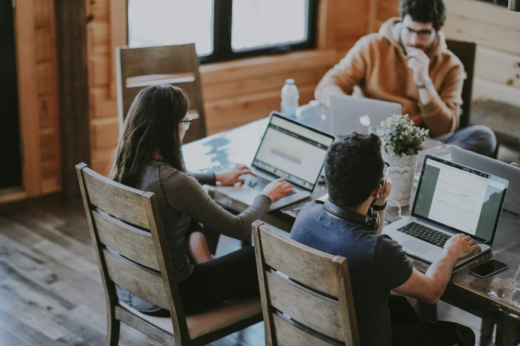 Three people work on laptops at a wooden table in a cozy room with wood-paneled walls.