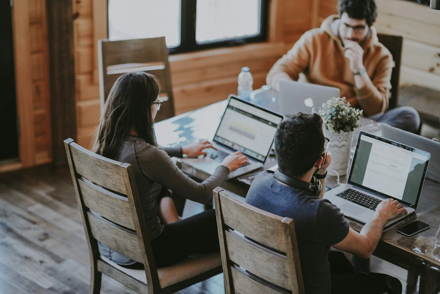 Three people work on laptops at a wooden table in a cozy room with wood-paneled walls.