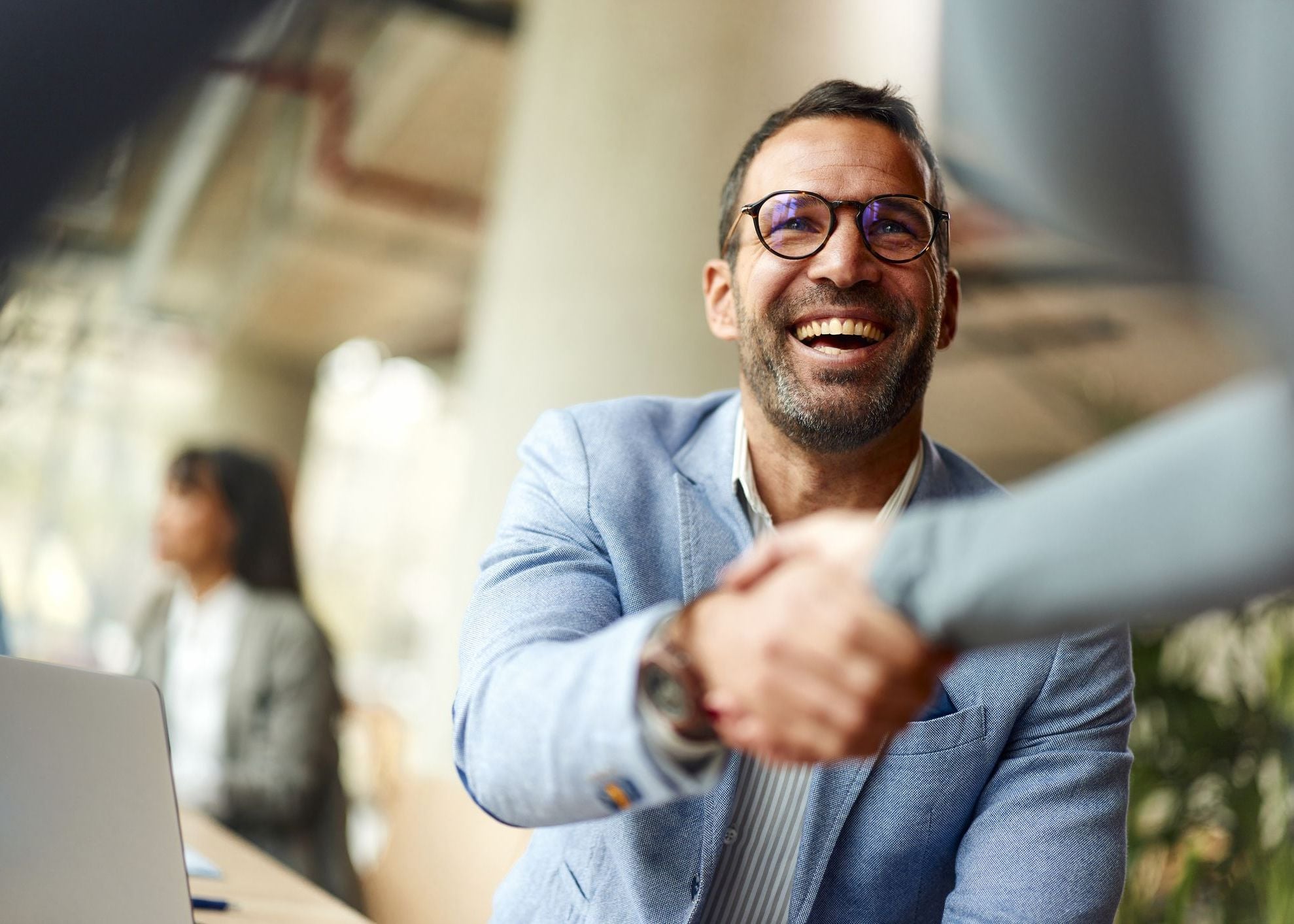 Smiling businessman in glasses shaking hands, with blurred office background and laptop.