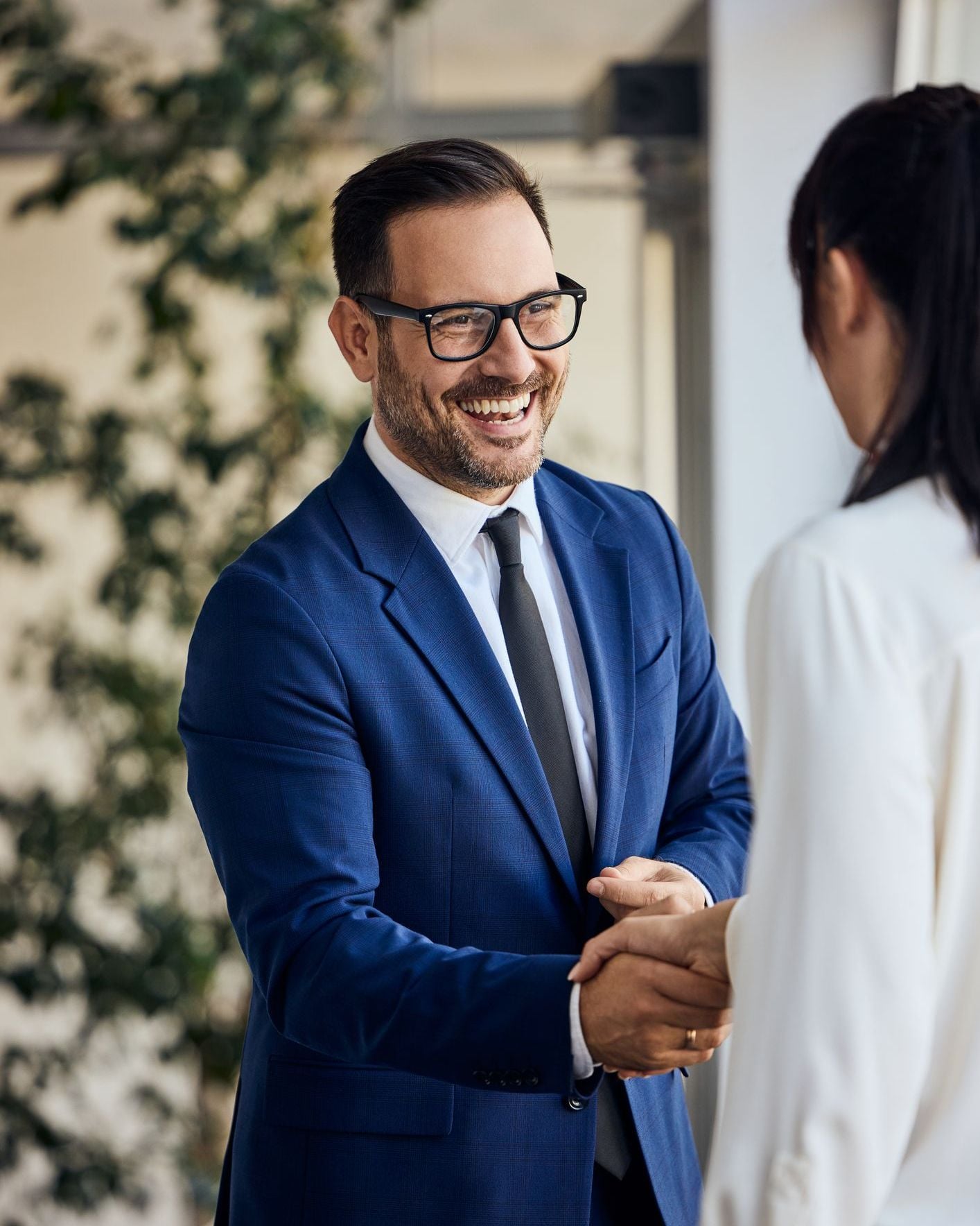Man in blue suit and glasses shaking hands with person in white shirt, smiling.