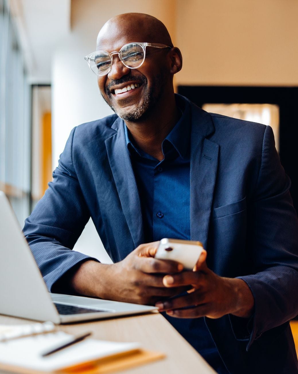 Smiling Black businessman in glasses and suit holding smartphone at desk with laptop.