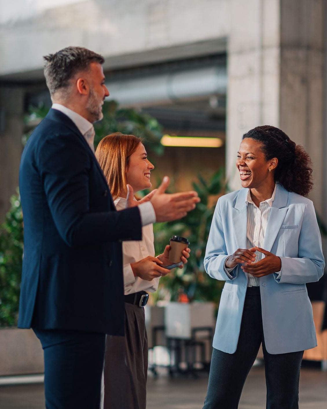 Three business professionals in a modern office, one man and two women, talking and laughing together.