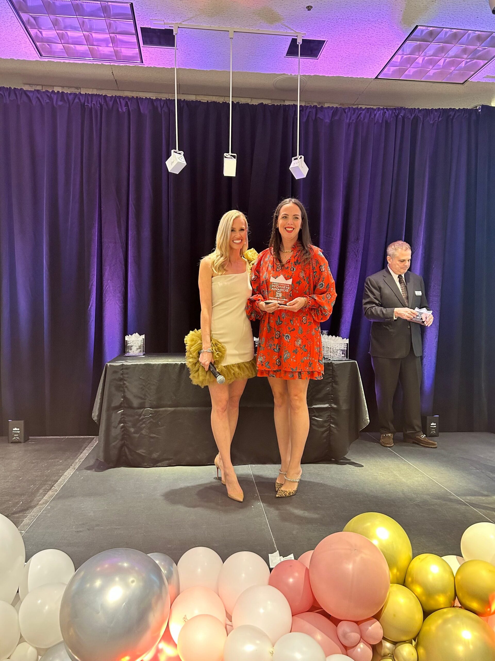 Two women holding awards on stage, one in a tan dress, other in red floral dress, with balloons and curtains.